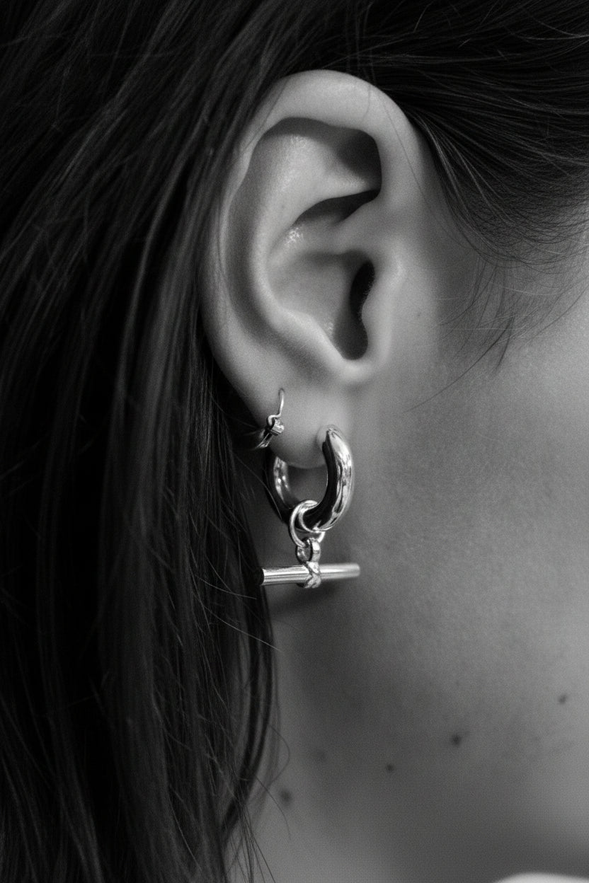 A close-up of a person's ear wearing a silver crescent hoop earring with a small toggle clasp, shown against dark brown hair.