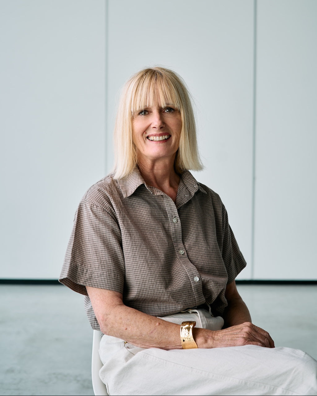 A smiling blonde woman seated indoors against a light gray paneled wall, wearing a short-sleeve brown patterned button-up shirt and white pants, with a gold watch visible on her wrist.