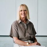 A smiling blonde woman seated indoors against a light gray paneled wall, wearing a short-sleeve brown patterned button-up shirt and white pants, with a gold watch visible on her wrist.