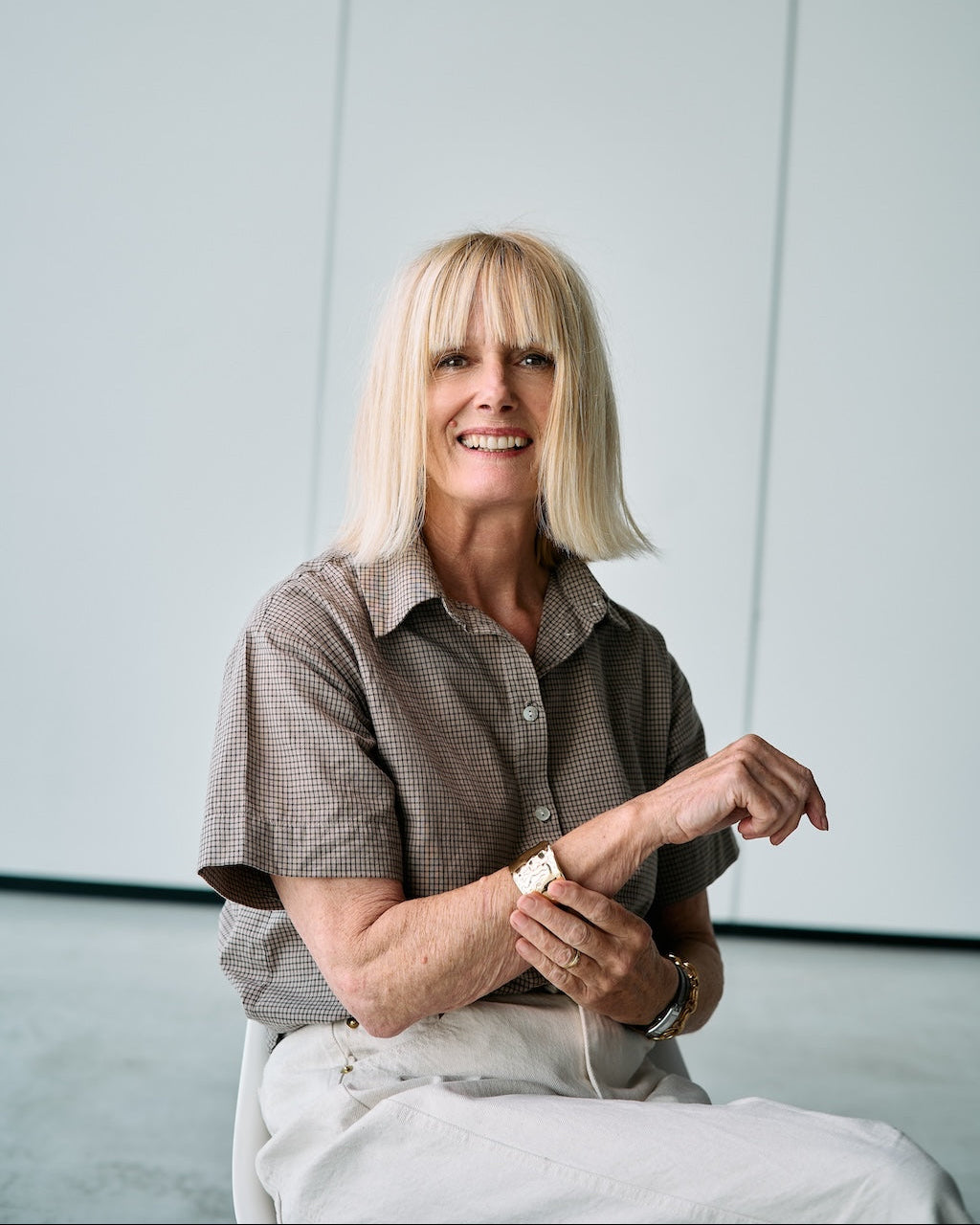 A smiling blonde woman seated against a light gray wall, wearing a brown short-sleeve button-up shirt and light beige pants.