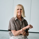 A smiling blonde woman seated against a light gray wall, wearing a brown short-sleeve button-up shirt and light beige pants.