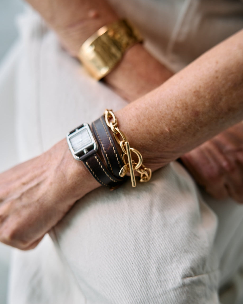 A close-up lifestyle shot of a person's wrist wearing multiple bracelets, including a gold chain bracelet, a gold toggle clasp bracelet, a dark brown leather strap bracelet, and a silver-toned watch, paired with light beige trousers.