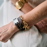 A close-up lifestyle shot of a person's wrist wearing multiple bracelets, including a gold chain bracelet, a gold toggle clasp bracelet, a dark brown leather strap bracelet, and a silver-toned watch, paired with light beige trousers.