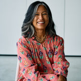 A smiling woman with dark gray hair sits wearing a pink long-sleeve blouse with a colorful floral pattern, layered gold necklaces, and hoop earrings against a minimalist light gray background.