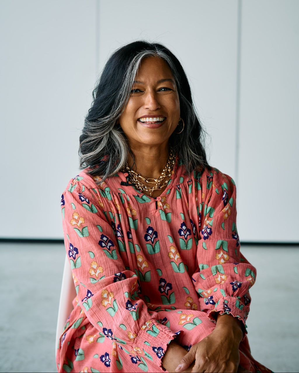A smiling woman with dark gray hair sits wearing a pink long-sleeve blouse with a colorful floral pattern, layered gold necklaces, and hoop earrings against a minimalist light gray background.