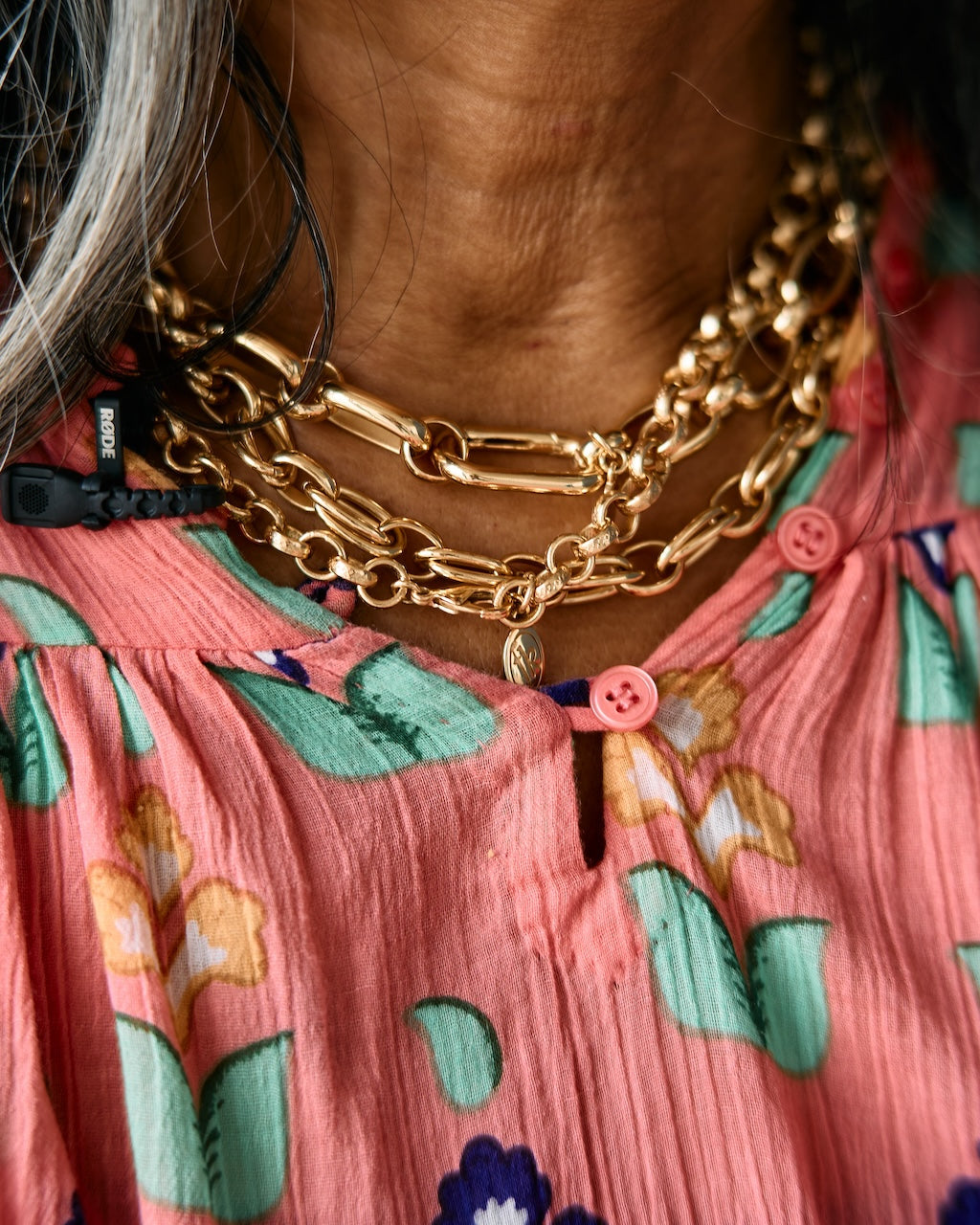 Close-up of a person wearing a coral pink floral top with green and blue flowers, layered gold chain necklaces, and a black cord necklace.