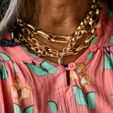 Close-up of a person wearing a coral pink floral top with green and blue flowers, layered gold chain necklaces, and a black cord necklace.
