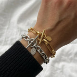 A close-up of a person's wrist wearing two chain bracelets, one gold and one silver, both with toggle clasp details and small round charms, against a white textured fabric background.
