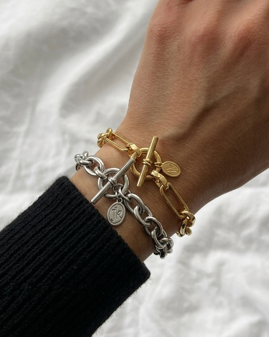 A close-up of a person's wrist wearing two chain bracelets, one gold and one silver, both with toggle clasp details and small round charms, against a white textured fabric background.