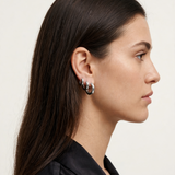 A close-up side profile of a woman with long dark brown hair wearing a pair of silver huggie hoop earrings, set against a plain off-white background.
