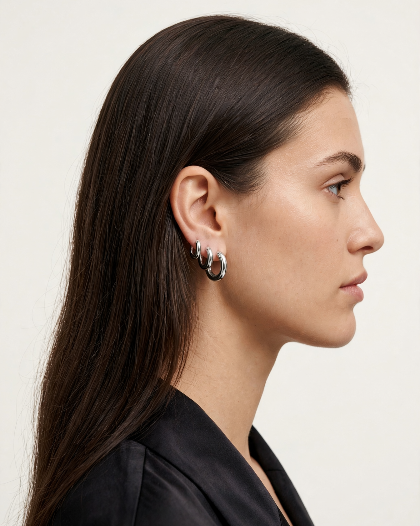A close-up side profile of a woman with long dark brown hair wearing a pair of silver huggie hoop earrings, set against a plain off-white background.