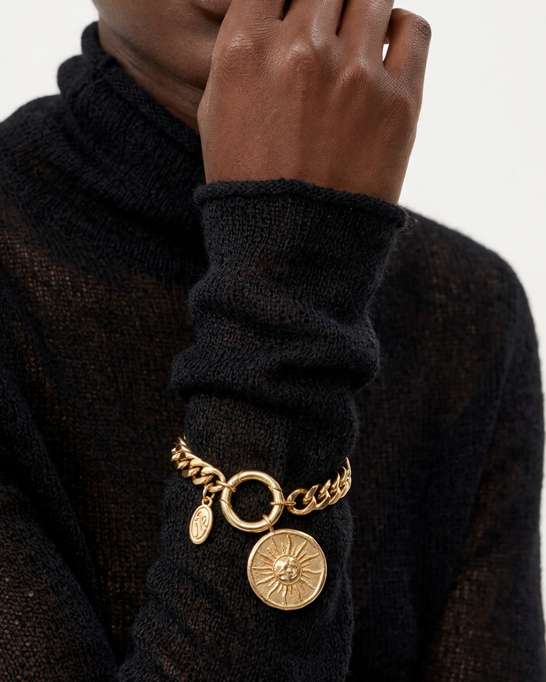 A close-up of a person wearing a black knit turtleneck sweater and a gold-tone chain bracelet with circular charms, including a sunburst medallion and a small oval tag, against a light gray background.