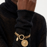 A close-up of a person wearing a black knit turtleneck sweater and a gold-tone chain bracelet with circular charms, including a sunburst medallion and a small oval tag, against a light gray background.